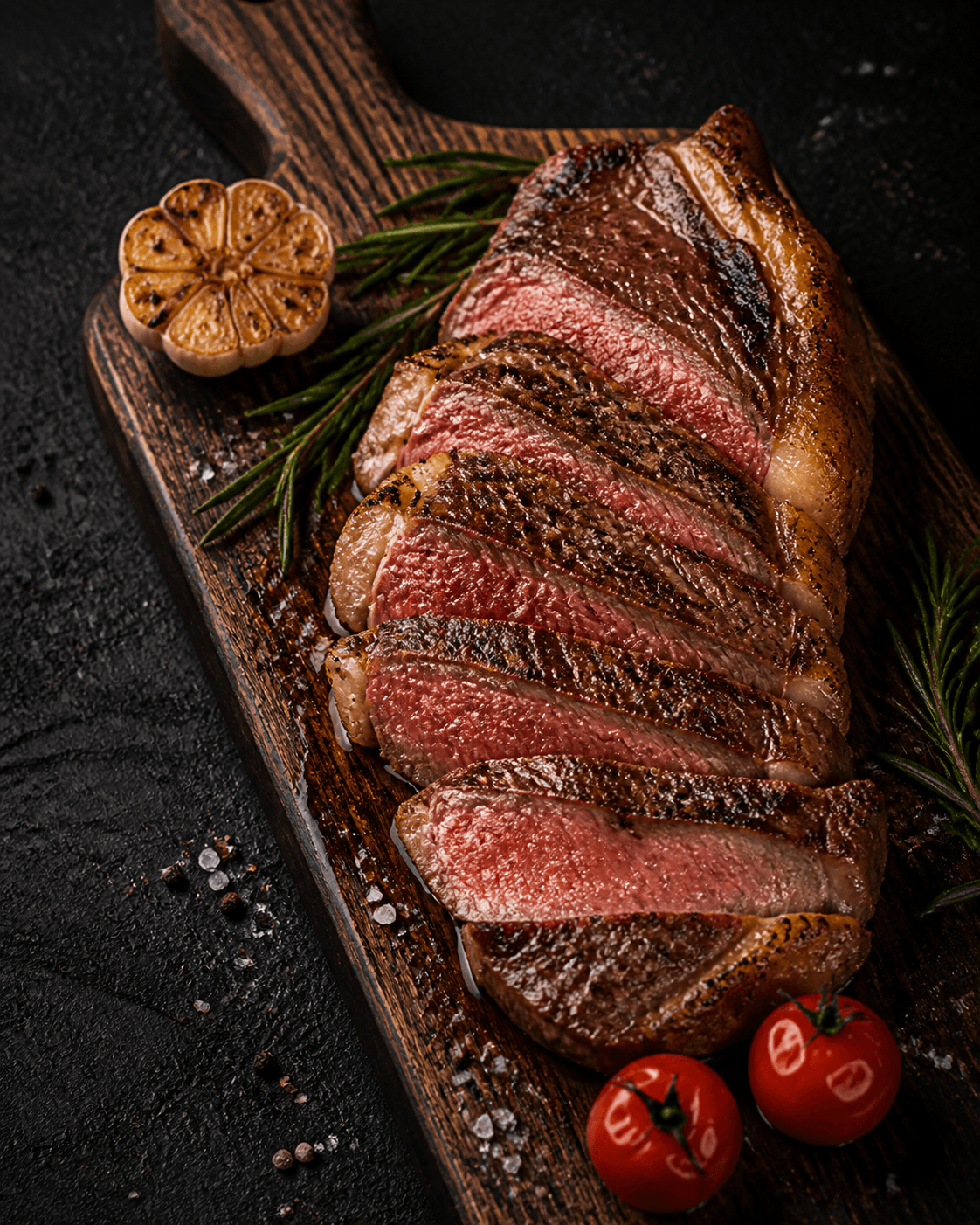 Corte de carne grelhado, fatiado, com crosta selada e interior rosado, servido sobre tábua de madeira com alecrim, alho assado e tomates